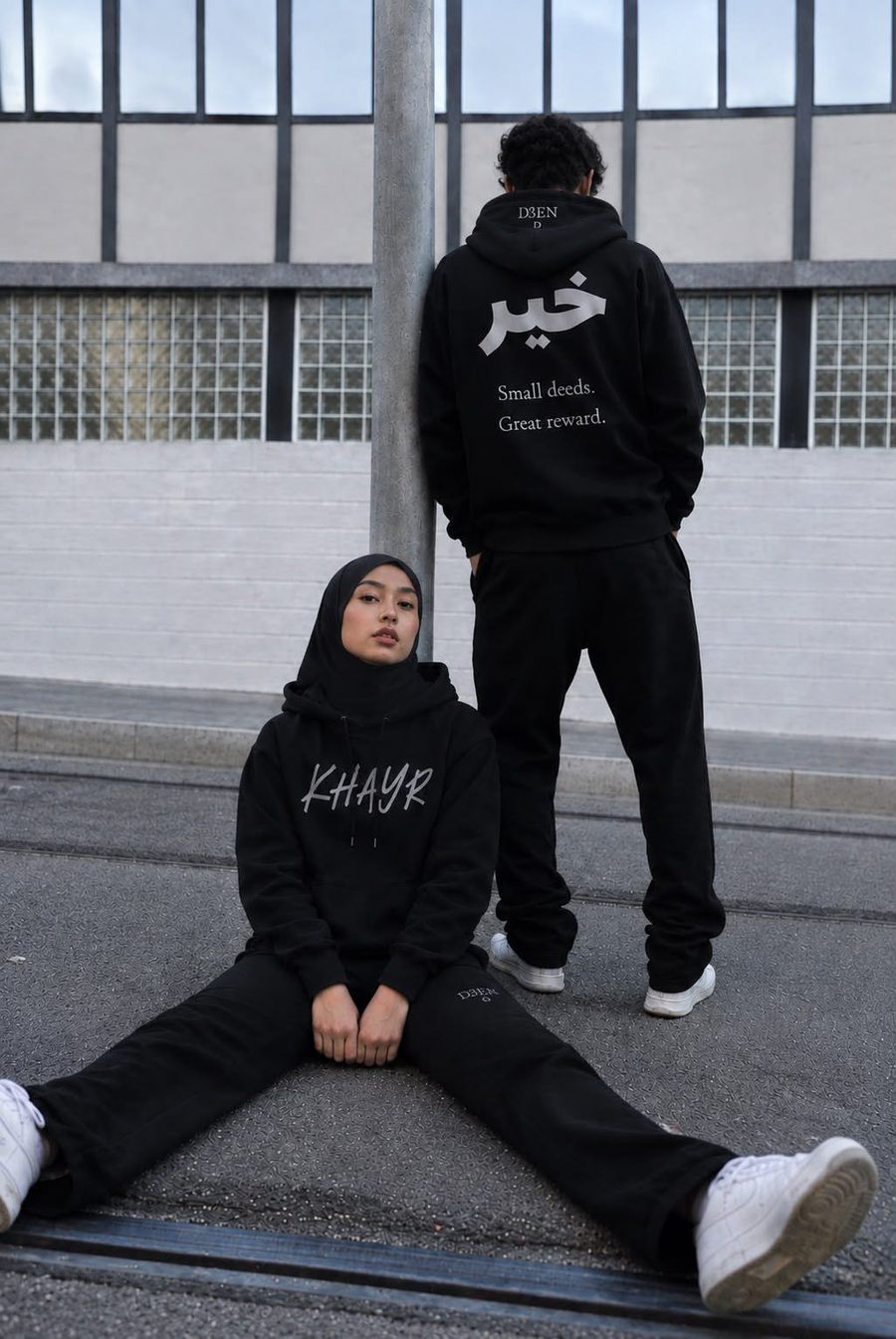 Two models in the D3EN Khayr hoodie and black trackpants, one seated on the street showing the front, one leaning on a pole showing the back